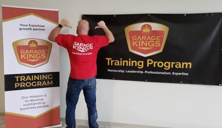 A person wearing a red "Garage Kings Certified Installer" shirt stands with their back to the camera, flexing their arms. They are in front of a black banner and a vertical banner, both displaying the "Garage Kings Training Program" logo and text.