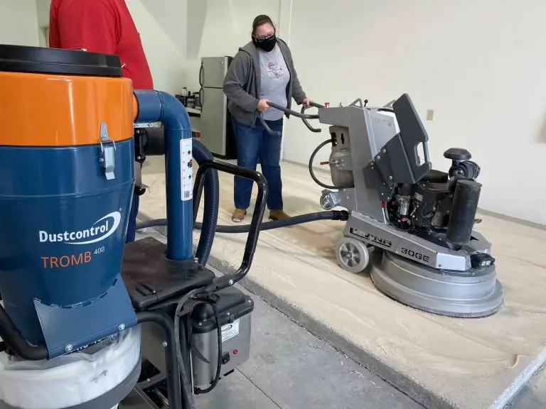 A masked person operates a large floor grinding machine on a concrete surface indoors. A Dustcontrol Tromb 400 vacuum system is visible in the foreground. Another person in a red shirt stands nearby, partially out of frame.