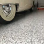 Close-up of a vintage car with white-walled tires parked on a speckled garage floor. The car's body is mostly obscured, showing just a portion of the door and the front wheel. There is a red toolbox in the background.