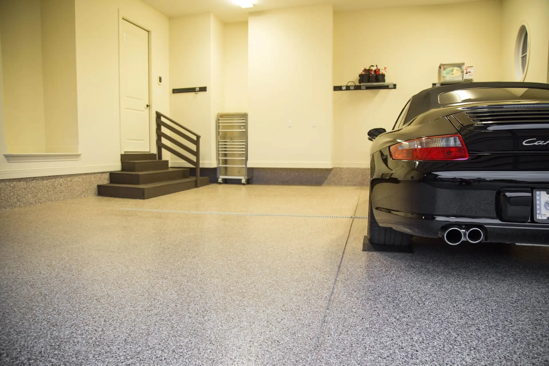A clean and modern garage with a black sports car parked inside. The garage has beige walls, a speckled floor, and a door leading to the house. Steps with a black railing lead up to the door, and shelves with various items are mounted on the wall.