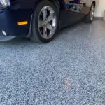 A black car with chrome wheels is parked on a polished speckled gray garage floor. The image shows the front-left corner of the car, the wheel, and the reflective surface of the floor. In the background are shelves with various items and a box against the wall.