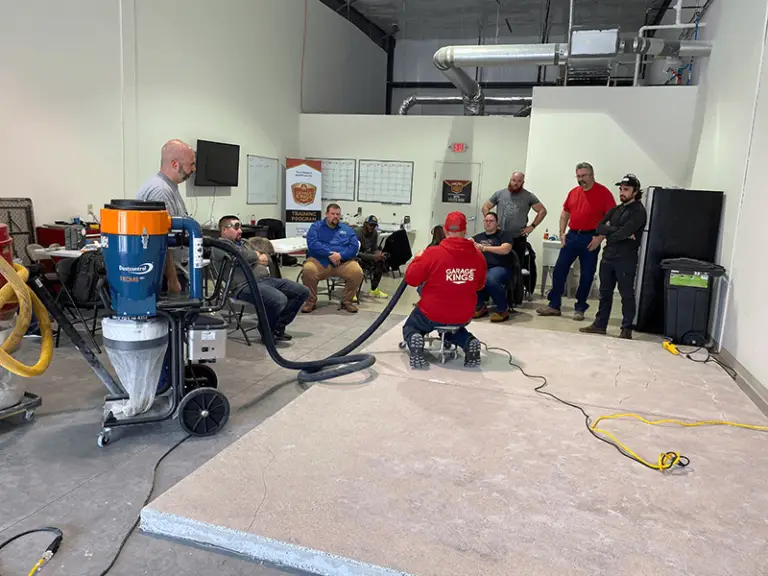 A group of men are gathered in a workshop setting. Two men in red shirts appear to be leading a demonstration with a large vacuum-like machine. Others are seated or standing, watching closely. The room is industrial with various equipment and a concrete floor.