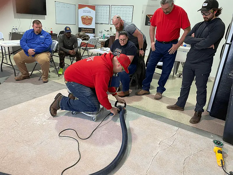 A man crouches on the floor using a tool on a large piece of carpet, surrounded by six attentive onlookers. The group appears to be part of a training session in an indoor setting with chairs, tables, and a screen in the background.