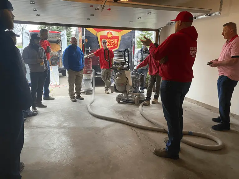 A group of people stand inside a garage, attentively surrounding a man in a red hoodie who is demonstrating how to use a floor polishing machine. A truck with "Garage Kings" branding is visible in the background through the open garage door.