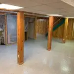 A basement with wooden paneling, support beams, and a dropped ceiling. The floor is covered with a speckled epoxy finish. There is a staircase with blue steps leading to an upper floor. Clear plastic sheets cover certain areas, possibly for renovations.