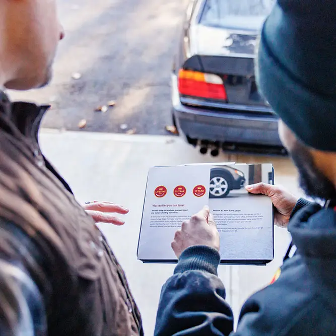 Two individuals are facing away from the camera, reviewing a brochure or document together. One appears to be pointing at images of a car on the page. They are standing near a driveway with a parked car in the background, and beside them is a garage filled with neatly organized storage cabinets.