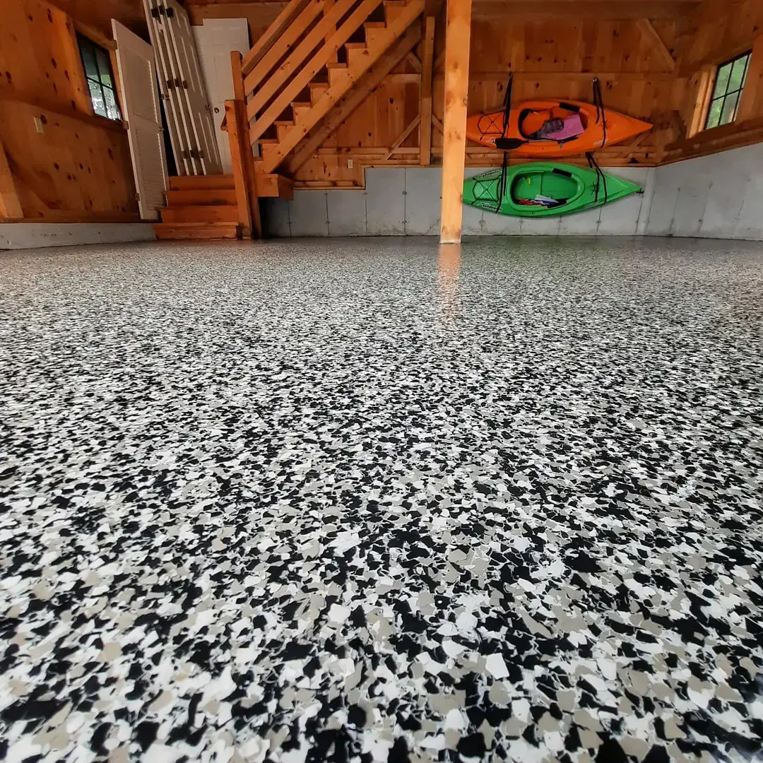 A garage with a speckled epoxy floor, enhanced by basement floor coatings, features a wooden staircase leading to an upper level. Two colorful kayaks, one orange and one green, are mounted on the far wall. Wood-paneled walls and windows are visible in the background.