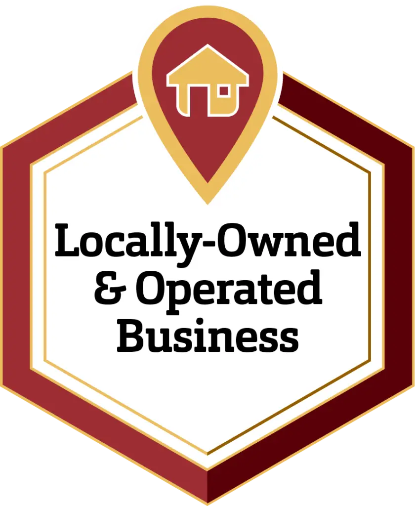 A hexagonal badge with a red and gold border features a house icon inside a map pin at the top. The text "Locally-Owned & Operated Business" is centered within the badge on a white background.