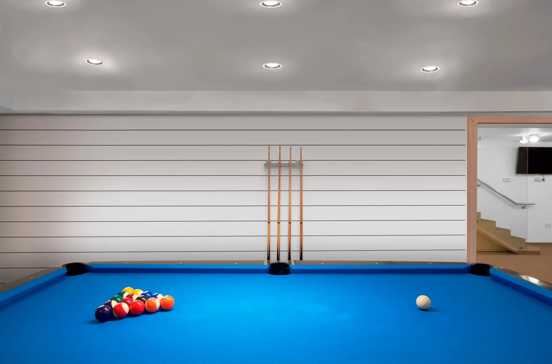 A modern, well-lit game room featuring a blue felt billiards table with a triangle of racked balls and a cue ball. Four pool cues are mounted on the wall in the background. Once just a storage space, this stunning area is the result of an impressive garage renovation, showcasing both style and functionality.