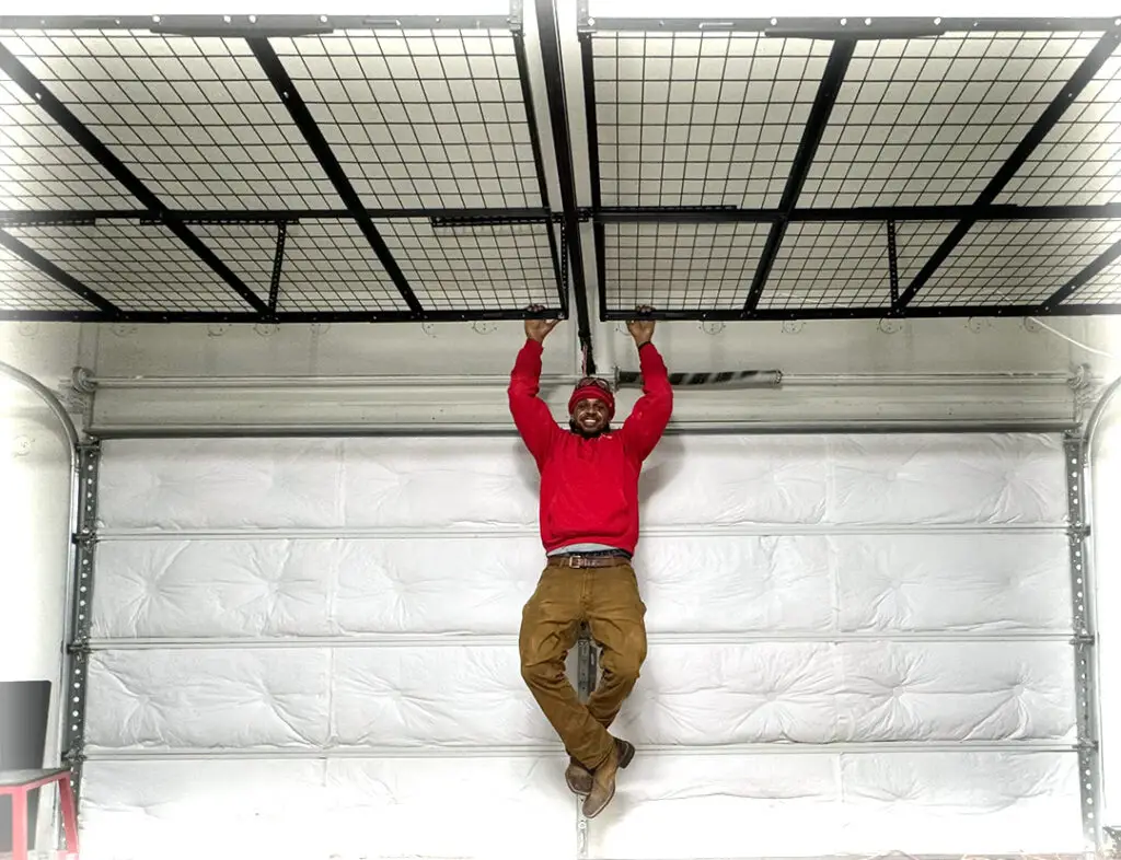 A man in a red sweater and brown pants is doing a pull-up while hanging from a ceiling-mounted metal rack in a garage. The garage door is closed and the walls are white. The man is looking at the camera and smiling.