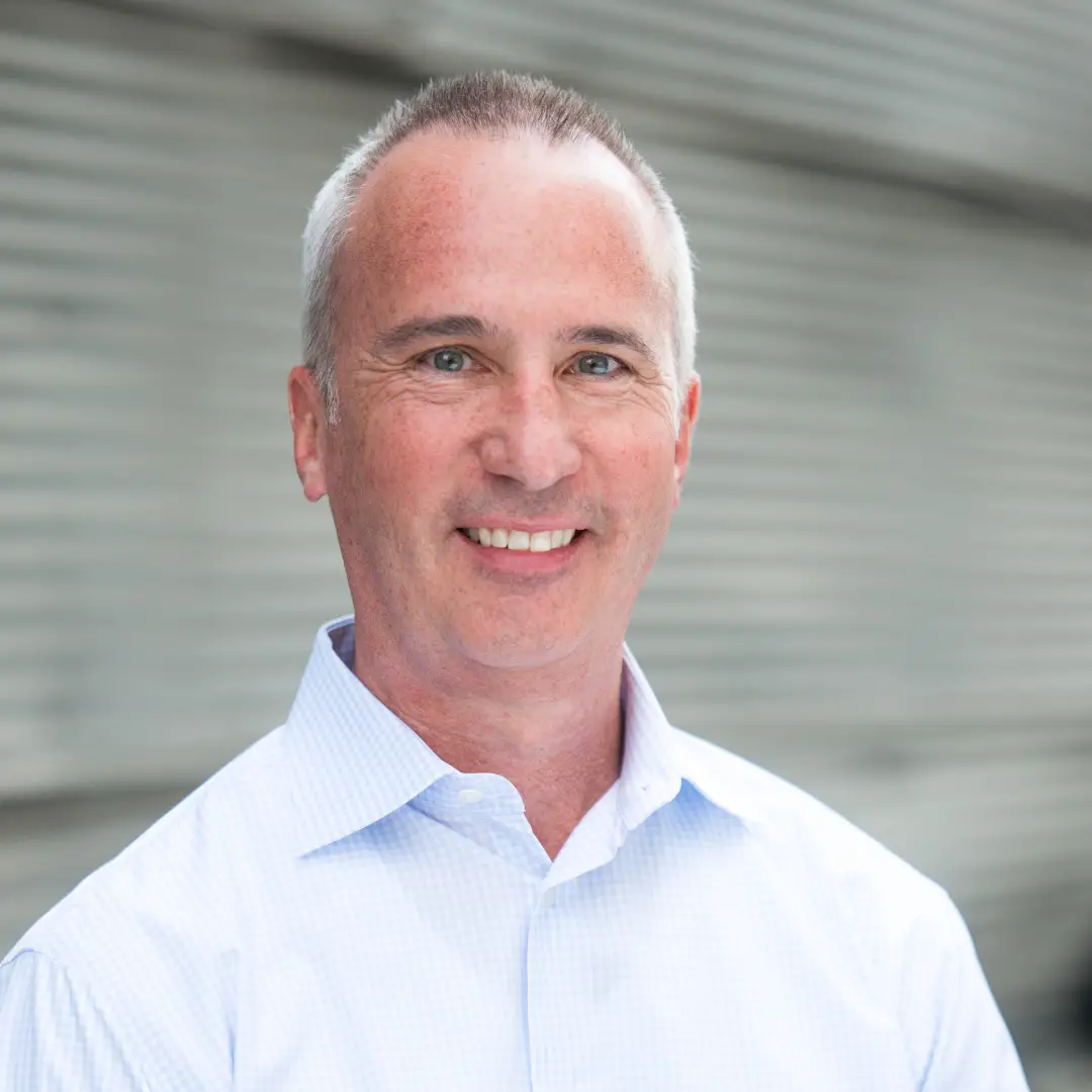 A smiling man with short gray hair is wearing a light blue collared shirt. The background is blurred, featuring horizontal gray lines.