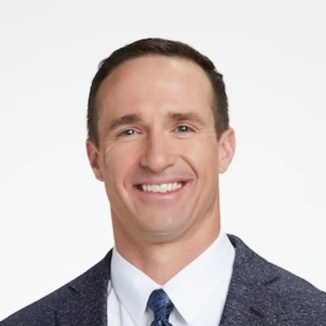 A man with short brown hair, wearing a white dress shirt, blue patterned tie, and dark blue suit jacket, is smiling in front of a plain white background.