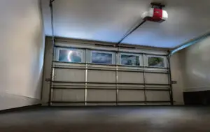 Interior view of an empty garage with a closed, segmented metal garage door. Through the windows, a storm with lightning is visible outside. An automatic garage door opener is attached to the ceiling. The garage is well-lit with smooth concrete flooring.