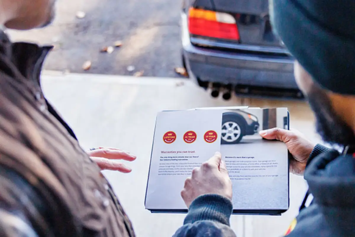 Two people stand next to a car. One person holds a clipboard with a document displaying car maintenance information, while the other person points to the document. Part of the car's rear is visible in the background. They appear to be discussing car maintenance.