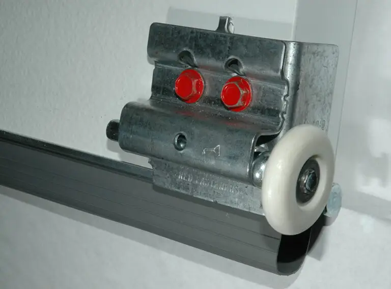 Close-up of a metal sliding mechanism, with two red buttons and a white plastic wheel attached to a metal track. The device appears to be mounted on a white surface, likely part of a door or drawer system.