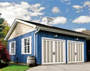 A quaint blue barn-style building with a stone facade near the roof and two large white garage doors. It has a small window beside each garage door and a partially cloudy sky in the background. A barrel planter is placed near the side window.