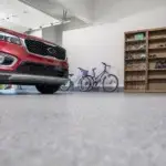 A clean garage with a red SUV parked inside. To the side, there are two bicycles leaning against the wall and a wooden shelf filled with various items, including helmets. The floor is spotless and has a smooth, grey finish.