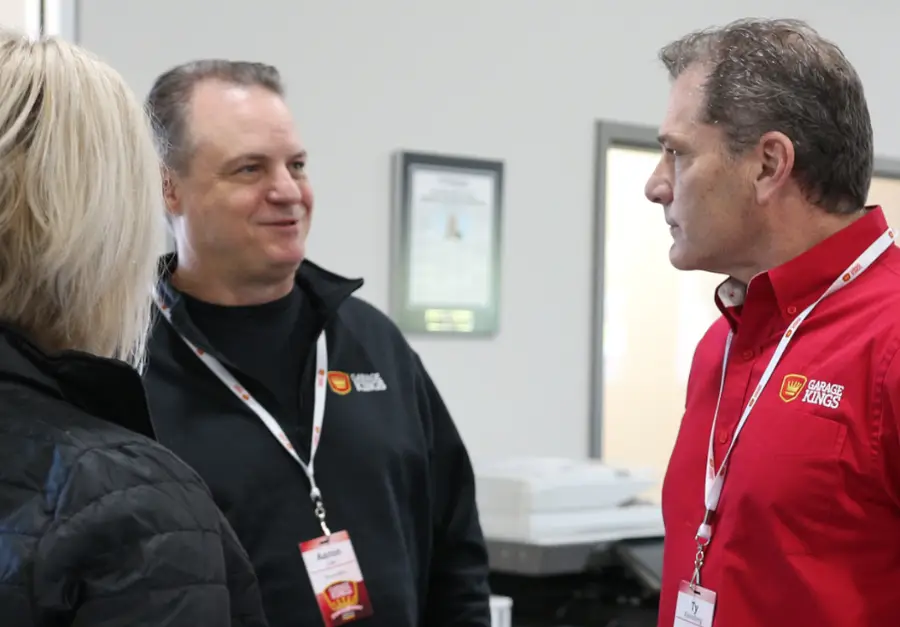 Three individuals are conversing indoors. Two men wearing lanyards, one in a black jacket and the other in a red shirt with a "Garage Kings" logo, face each other. A woman in a black jacket with short blonde hair stands with her back to the camera.