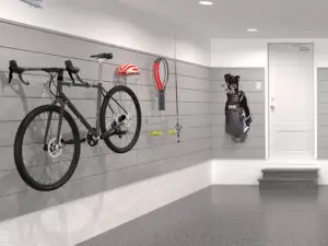 Well-organized garage with a bicycle mounted on the left wall, a red helmet, and various tools hanging nearby. Golf clubs rest against the same wall near a door with steps leading up to it. The floor is clean and gray, and the walls are painted light gray.