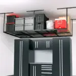 A garage storage system featuring a ceiling-mounted shelving unit with various items stored, including plastic bins, a red cooler, and a paint bucket. Below, there are sleek, black storage cabinets with silver handles and drawers, providing ample storage space.