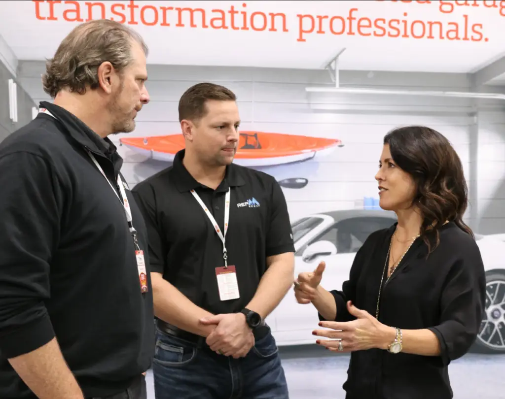 Three people, two men and one woman, engage in conversation at an indoor event. The background features a sign reading "transformation professionals" and a car in a garage setup. They are wearing lanyards with IDs, and the woman is gesturing with her hands.