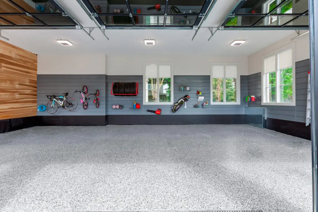 A clean, organized garage with grey walls and a speckled floor. Bicycles, tools, and other equipment are neatly mounted on the walls. Large windows let in natural light, and an elevated storage rack hangs from the ceiling. The space is tidy and well-maintained.