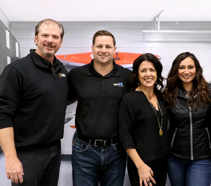 Four people are posing indoors, standing side by side and smiling at the camera. The two men on the left are wearing black tops, while the two women on the right are dressed in dark-colored clothing with one wearing a necklace. The background appears to be a garage or workshop with visible equipment.