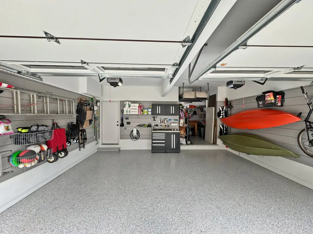 A well-organized garage with various tools and equipment neatly stored on wall-mounted shelves and racks. Two kayaks are hung on the right wall. The garage floor is clean and speckled, and there is minimal clutter, showcasing an efficient storage system.