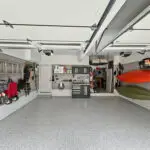 A well-organized garage with various tools and equipment neatly stored on wall-mounted shelves and racks. Two kayaks are hung on the right wall. The garage floor is clean and speckled, and there is minimal clutter, showcasing an efficient storage system.