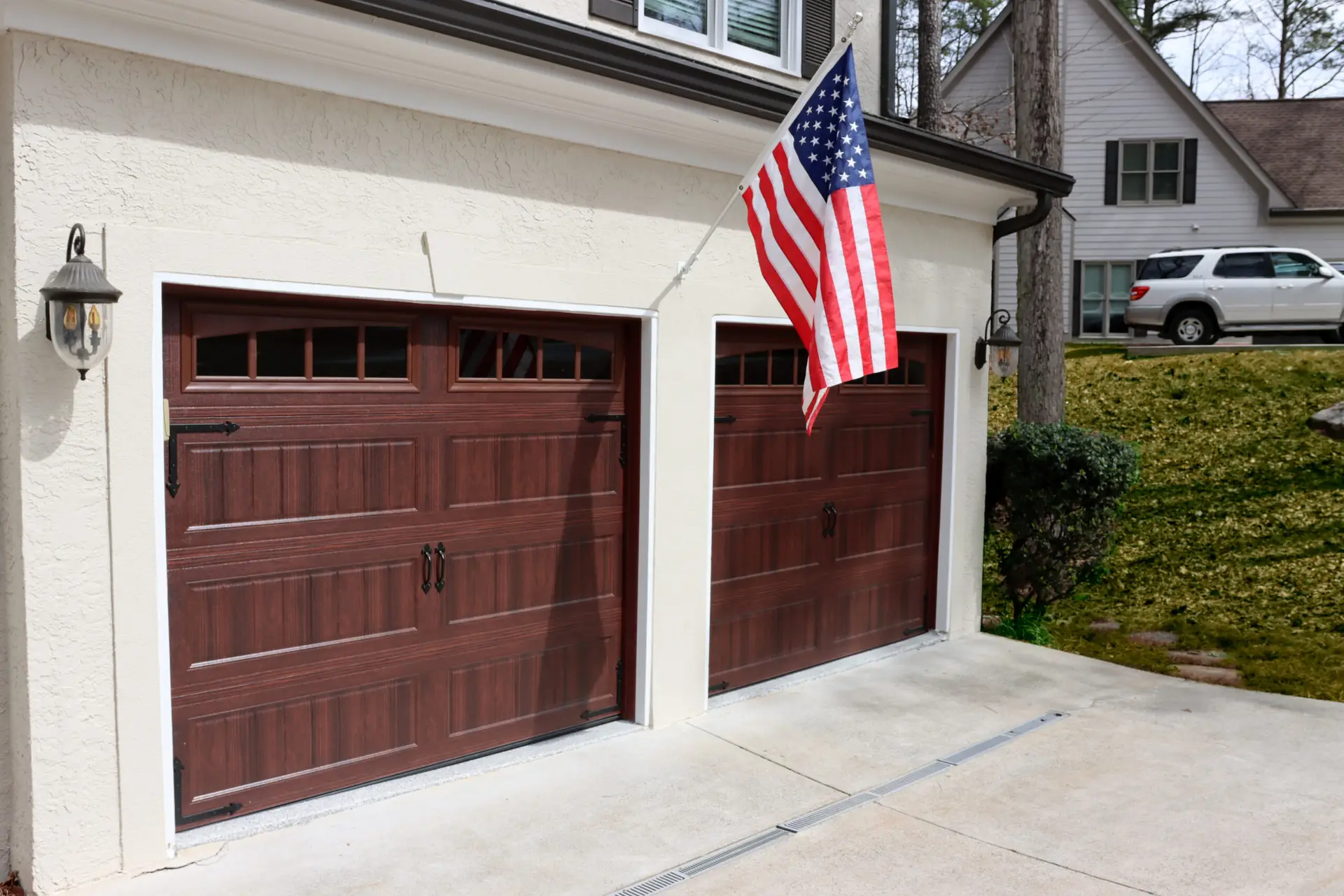 GK-Garage-Doors-US-Flag-02 | Garage Kings A house with two dark brown garage doors, an American flag mounted on the exterior wall between them, and a small lamp hanging above the left garage door. There's a white SUV parked on the driveway of the adjacent house, and trees are visible in the background.