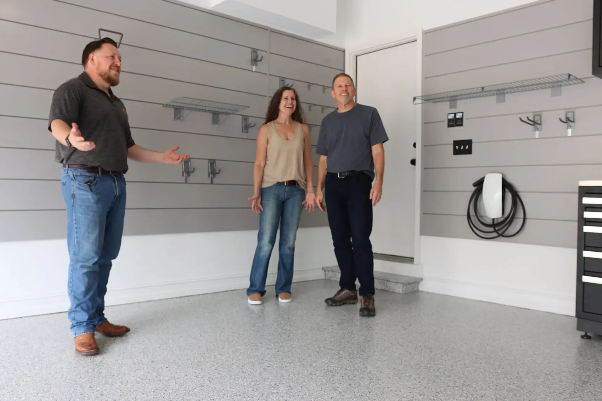 Three people stand in a garage with gray walls and organized shelving. On the left, a person gestures while speaking, and two people in casual clothing stand in the center. The garage features a clean, epoxy-coated floor and various mounted hooks and shelves.