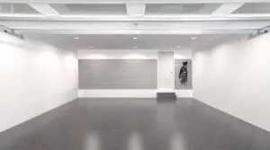 An empty, spotless garage with polished gray epoxy flooring and white walls. A section of the right wall has horizontal shelving. There is a white door with silver handle leading out, and the ceiling features recessed lighting.