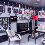 A stylish lounge area with black and gray decor, featuring a widescreen TV surrounded by shelves filled with various liquor bottles and decorative items. The room has a beige sofa set, a glass coffee table, and a "Make Your Move" poster on the wall.