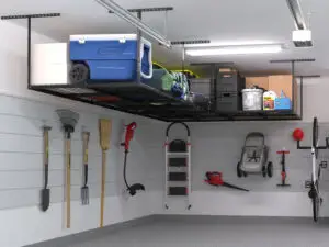 A well-organized garage with a ceiling storage rack holding various items including a blue cooler, green and black bins, and a gas can. Wall-mounted tools like shovels, rakes, a broom, and a leaf blower are neatly hung below. A bicycle and a stroller are also stored.