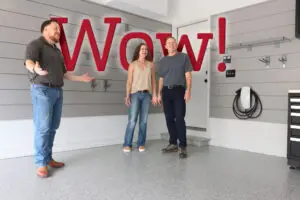 Three people stand in an organized garage with a clean, finished floor and new mounted storage units. One person gestures animatedly, while the other two are looking around. The word "Wow!" is overlaid in big red letters, capturing their excitement at the offer.