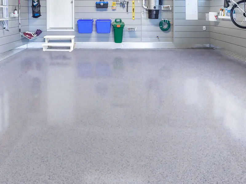 A clean, organized garage features shiny, speckled polyaspartic garage flooring, storage bins, hanging tools, shelves, a bike, and a green hose on the wall. White steps lead up to a door on the left side.