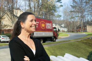 A woman stands outdoors with a smile and arms crossed, as a "Garage Kings" truck is parked on a tree-lined suburban street. The sunny February day highlights the scene's charm, offering a glimpse into serene community life.