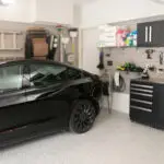 A sleek black car is parked in a well-organized garage, embodying why 2025 is the year to finally love your garage. It features wall-mounted ladders, tools, and cleaning supplies alongside two black storage cabinets with work surfaces. The speckled floor enhances the pristine atmosphere.