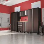 A neat garage with red walls featuring a set of sleek black cabinets and drawers. The workspace is organized, with overhead lighting, slat walls for storage, and a clear, glossy floor. A small window allows natural light to enter.