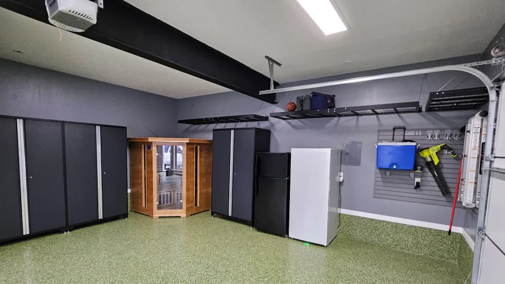A spacious, organized garage with green speckled flooring features the best garage storage system—tall black cabinets, a wooden sauna, a mini fridge, wall-mounted shelves holding bins and bags, plus a blue cooler on a gray slat wall.