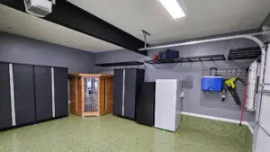 A clean garage interior showcasing dark cabinets, a small wooden storage unit, and a white refrigerator. Shelving organizes various items, including a bag. A blue cooler and gardening tools hang on the wall above a green-speckled floor—a testament to mastering Garage Organization amid March madness.