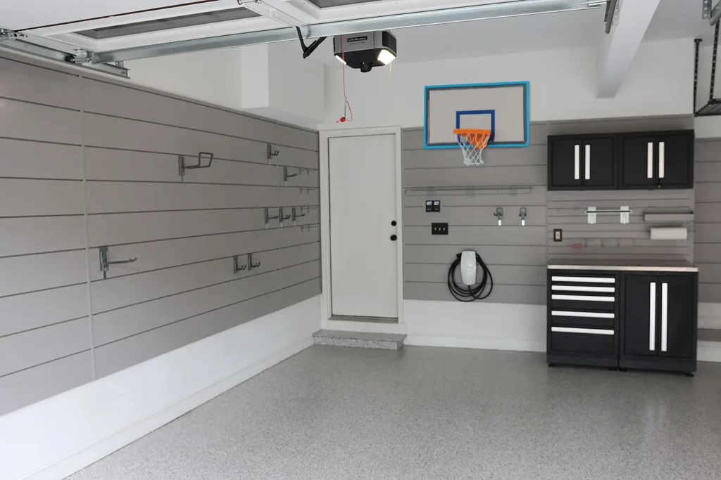 A clean, organized garage with gray slat walls, hooks for storage, a small basketball hoop above the door, a charging station, black storage cabinets with a wooden countertop, and a speckled light gray floor showcases smart garage organization.