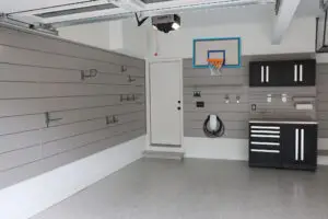 Organized garage interior featuring gray slat walls with hooks and shelves for efficient garage organization. A black cabinet with silver handles, white charging unit, small basketball hoop above the door, and clean speckled floor complete this March-ready setup.