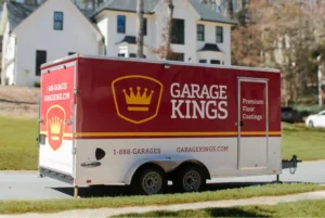 A red and white Garage Kings trailer is parked on a suburban street, featuring a gold crown logo and text advertising premium floor coatings, a phone number, and a website. Houses and trees are visible in the background.