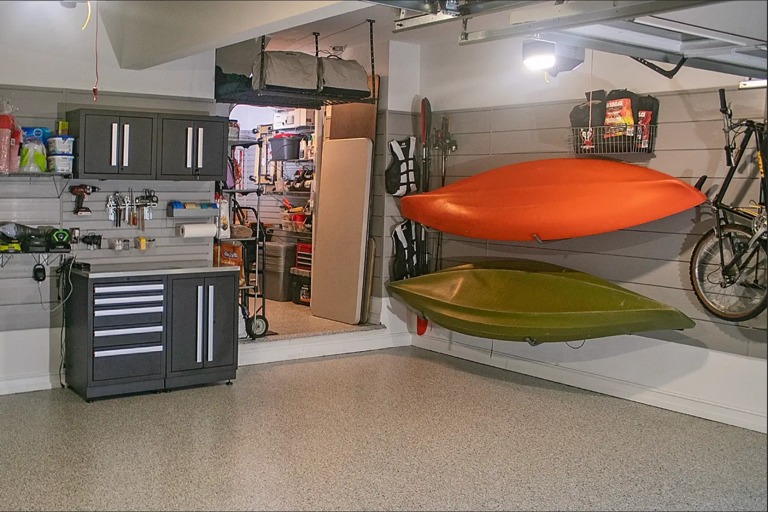 A clean, organized garage showcases a stunning garage renovation with storage cabinets, a workbench, and two kayaks—one orange, one green—mounted on the wall. Bicycles and shelves line the space, while an open door leads to more storage.