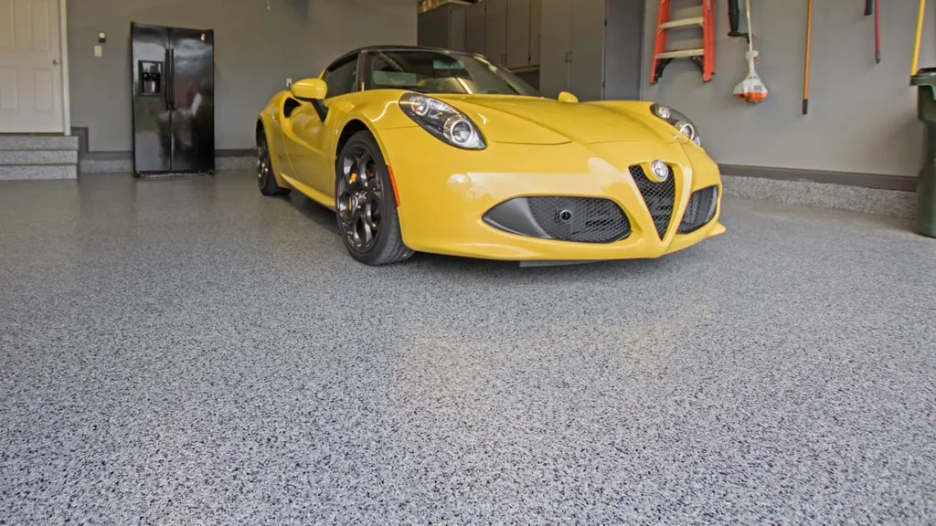 A yellow sports car is parked in a clean, modern garage with a speckled polyaspartic floor coating. Cabinets, a ladder, and some tools are mounted on the gray walls in the background.