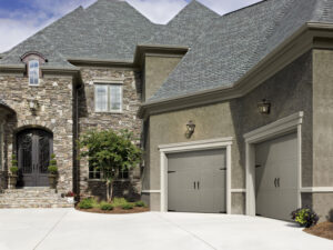 A large, elegant house with stone and stucco exterior, arched black front door, two gray garage doors, manicured shrubs, and a paved driveway on a sunny day.
