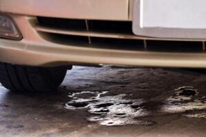 A close-up of the front of a beige car parked on a concrete surface with a noticeable dark fluid leak underneath the vehicle—highlighting how Garage floor coatings can help protect against stains in wet conditions.