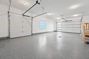 Spacious, clean, and empty three-car garage with gray speckled flooring—a perfect upgrade featured in our 2025 Gift Guide. White walls, bright ceiling lights, a window, and a wooden staircase on the right complete the look.