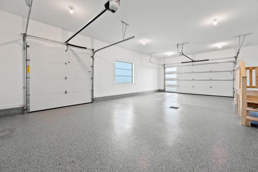 Spacious, clean, and empty three-car garage with gray speckled flooring—a perfect upgrade featured in our 2025 Gift Guide. White walls, bright ceiling lights, a window, and a wooden staircase on the right complete the look.